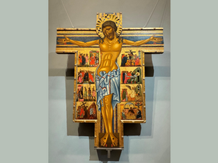 Crucifix with a golden Jesus, arms outstretched, surrounded by colorful religious scenes on a wooden cross, set against a gray wall.