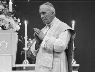 Bishop in white vestments stands in church, hands clasped in prayer beside lit candles and flowers, solemn black-and-white scene