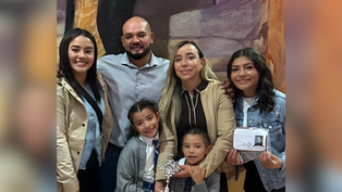 Family of six smiling in front of a colorful mural. One holds a trophy, another a photo ID. Casual attire, warm atmosphere.