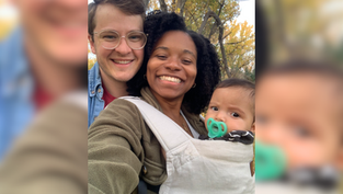 Smiling couple with a baby in a carrier outdoors, surrounded by autumn trees. The baby has a pacifier. Bright and joyful scene.