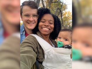 Smiling couple with a baby in a carrier outdoors, surrounded by autumn trees. The baby has a pacifier. Bright and joyful scene.