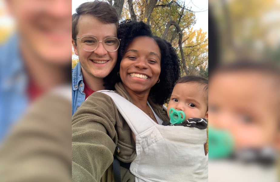 Smiling couple with a baby in a carrier outdoors, surrounded by autumn trees. The baby has a pacifier. Bright and joyful scene.