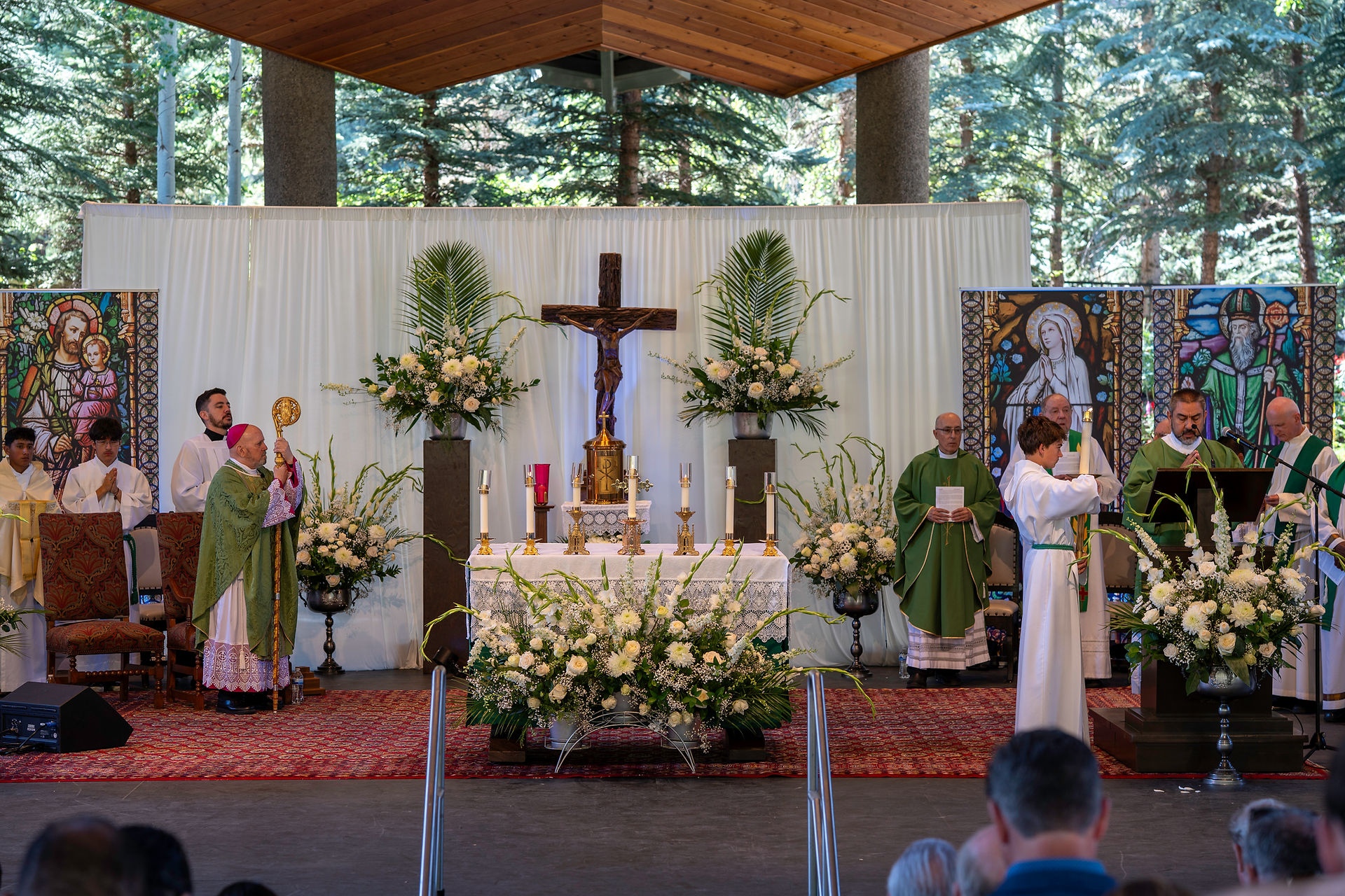 A religious ceremony in a church or chapel, featuring an altar with flowers, candles, and a crucifix. Clergy in colorful vestments stand near the altar, with stained glass and trees visible in the background. Attendees are seated in the foreground.