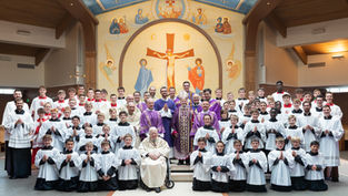 Group photo in a church with clergy, altar servers in white robes, and boys in red jackets. Iconic mural and crosses in the background.