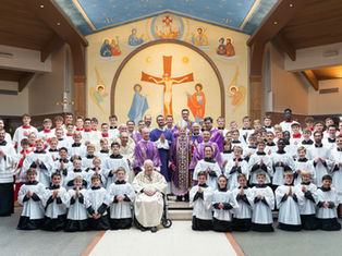 Group photo in a church with clergy, altar servers in white robes, and boys in red jackets. Iconic mural and crosses in the background.