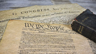 Historic documents with "We the People" and "In Congress" visible, next to an aged Holy Bible on a wooden table. Earthy tones.