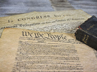 Historic documents with "We the People" and "In Congress" visible, next to an aged Holy Bible on a wooden table. Earthy tones.