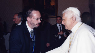 Two men shake hands, one in white religious attire with gold chains. Background has people and a framed painting, creating a formal setting.