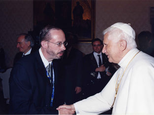 Two men shake hands, one in white religious attire with gold chains. Background has people and a framed painting, creating a formal setting.