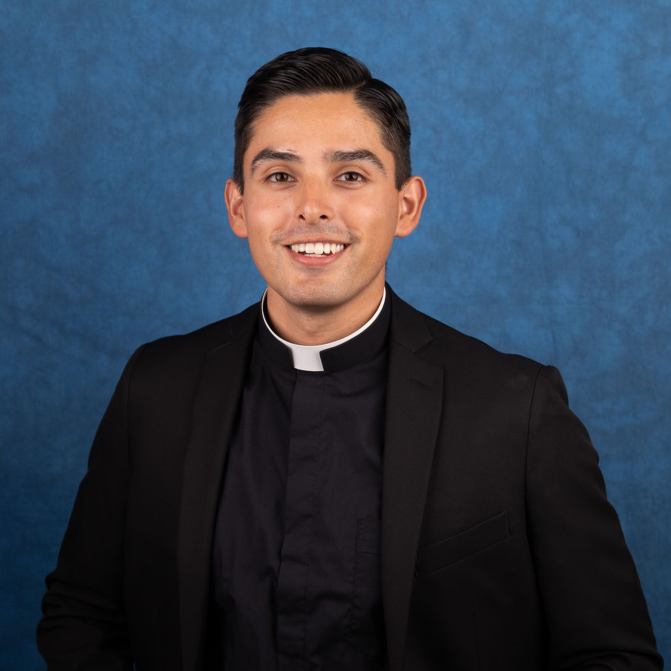 Smiling young priest in a black suit and clerical collar against a blue studio backdrop.