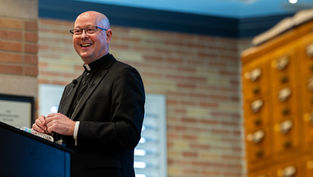 Clergyman in black attire smiles while speaking at a podium. Background shows a brick wall and wooden drawers in a warmly lit room.