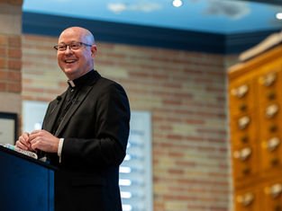 Clergyman in black attire smiles while speaking at a podium. Background shows a brick wall and wooden drawers in a warmly lit room.