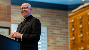 Clergyman in black attire smiles while speaking at a podium. Background shows a brick wall and wooden drawers in a warmly lit room.