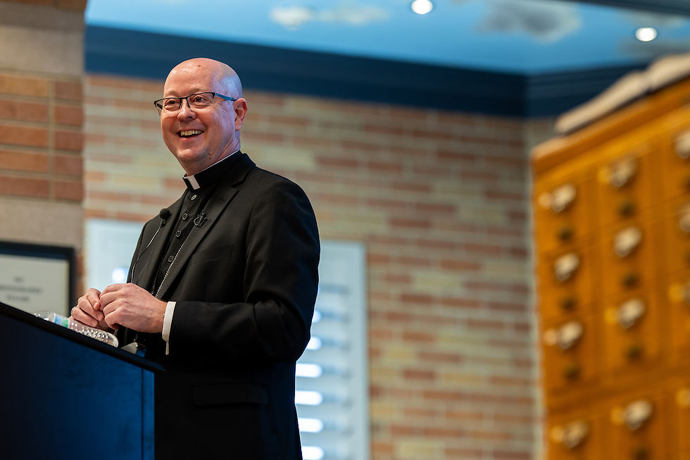 Clergyman in black attire smiles while speaking at a podium. Background shows a brick wall and wooden drawers in a warmly lit room.