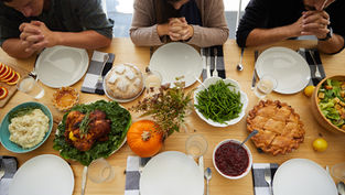 Three people with hands clasped in prayer around a table set with Thanksgiving foods like turkey, pie, and salad. Warm, inviting atmosphere.