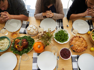 Three people with hands clasped in prayer around a table set with Thanksgiving foods like turkey, pie, and salad. Warm, inviting atmosphere.