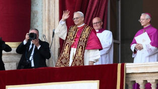 A man in ornate religious attire waves from a balcony, flanked by clerics in white and purple. A photographer captures the moment.