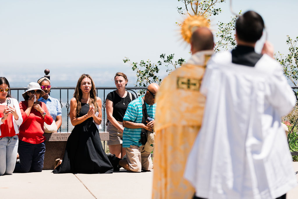People kneel in prayer at an outdoor Catholic service, with a priest in white and gold robes in the foreground.