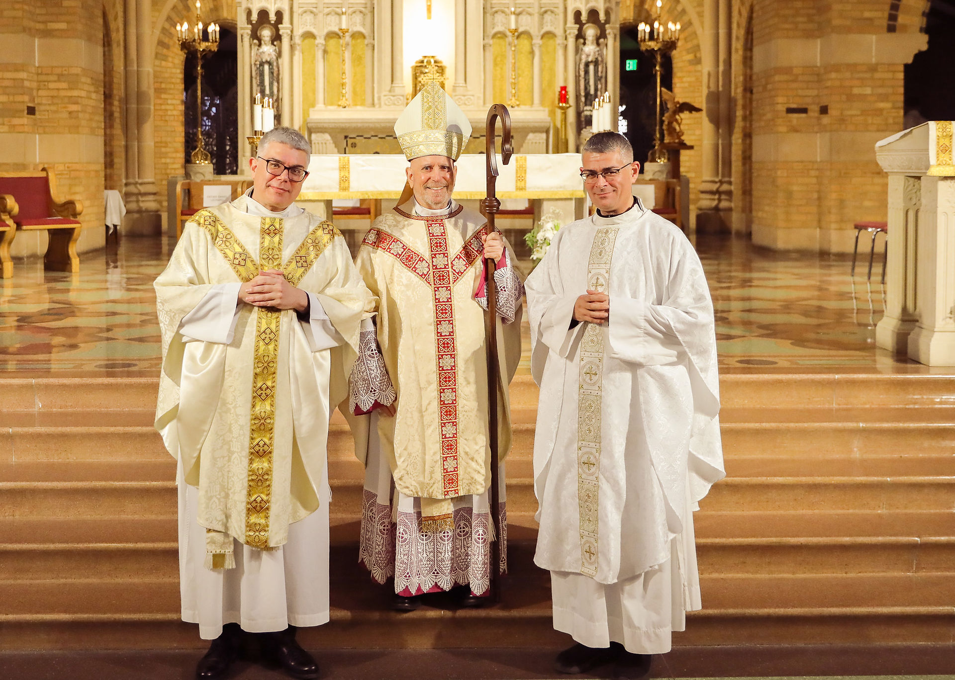 Two priests, who are also brothers, stand in an ornate chapel for a photo with an archbishop following a religious ceremony. The mood is celebratory.