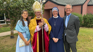 Four people smiling outside a brick building: a woman in blue, a bishop in gold and red attire, a nun in navy, and a man in a suit.