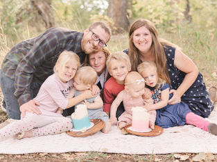 A family of seven smiling in a grassy setting. Two cakes with blue and pink frosting sit on plates. Warm, joyful atmosphere.
