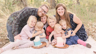 A family of seven smiling in a grassy setting. Two cakes with blue and pink frosting sit on plates. Warm, joyful atmosphere.