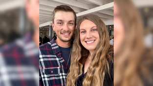 Smiling man and woman pose indoors. Man in plaid shirt, woman with long wavy hair. White ceiling beams in the background. Bright, happy mood.