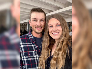 Smiling man and woman pose indoors. Man in plaid shirt, woman with long wavy hair. White ceiling beams in the background. Bright, happy mood.
