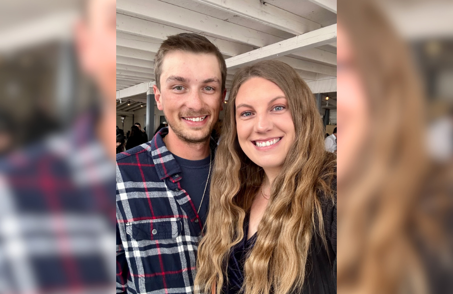 Smiling man and woman pose indoors. Man in plaid shirt, woman with long wavy hair. White ceiling beams in the background. Bright, happy mood.