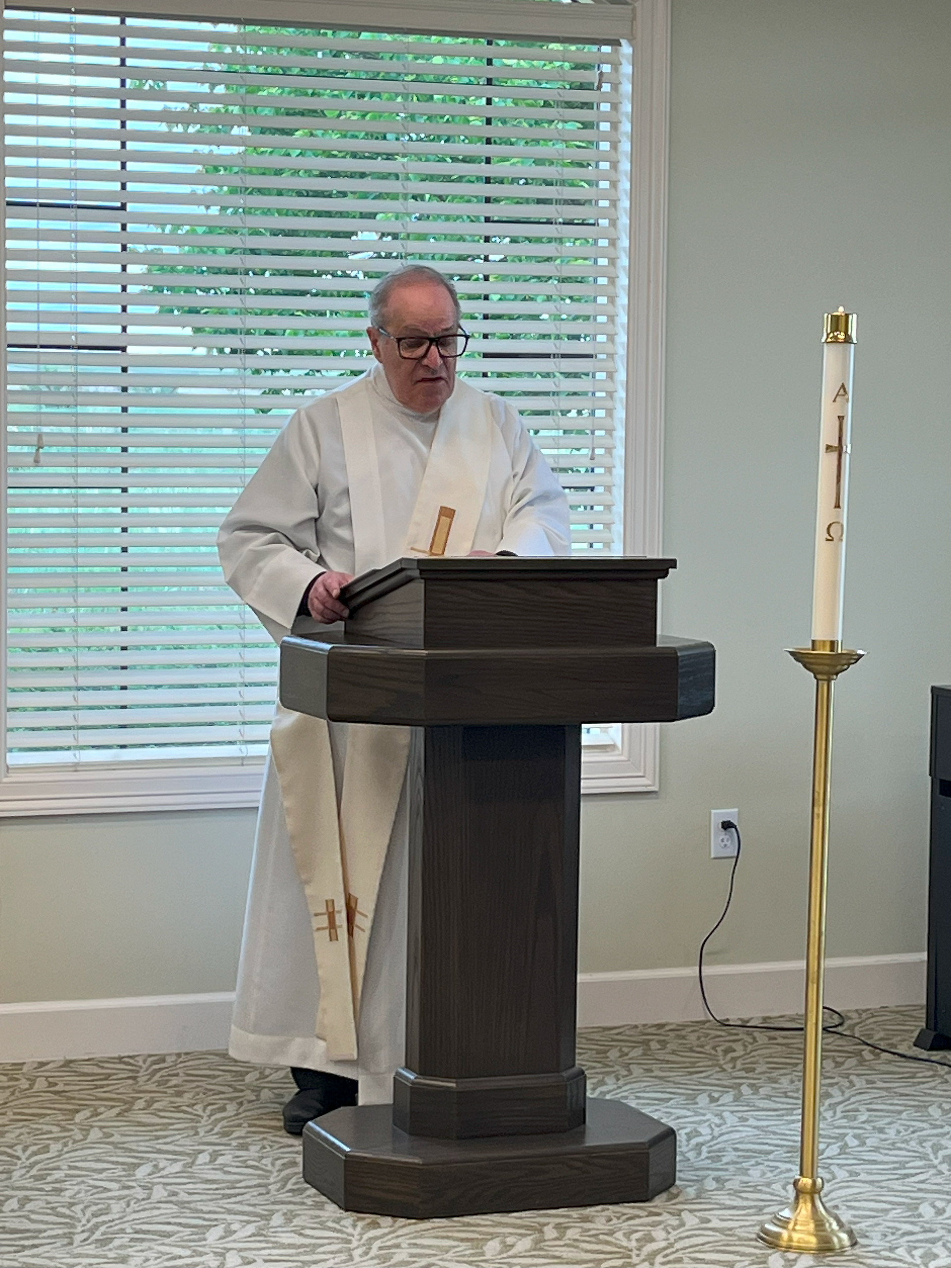 Clergy member in white robe speaks at a wooden lectern. A tall candle stands nearby. Bright room with blinds in the background.