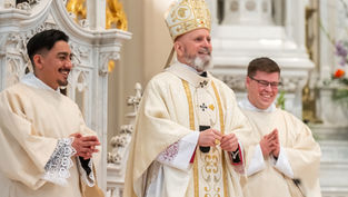 Three clergymen in ornate robes stand in a church, smiling. The central figure wears a detailed mitre. Decorative white and gold background.