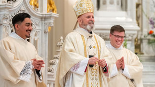 Three clergymen in ornate robes stand in a church, smiling. The central figure wears a detailed mitre. Decorative white and gold background.
