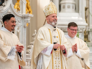 Three clergymen in ornate robes stand in a church, smiling. The central figure wears a detailed mitre. Decorative white and gold background.