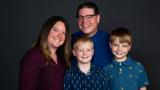 Smiling family of four posing against a dark background. Adults wear maroon and blue shirts; children in patterned blue shirts.