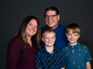 Smiling family of four posing against a dark background. Adults wear maroon and blue shirts; children in patterned blue shirts.