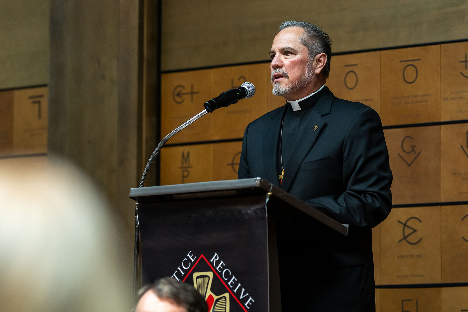 A Catholic deacon in a black clerical suit speaks at a podium with a microphone, addressing an audience in a formal indoor setting.