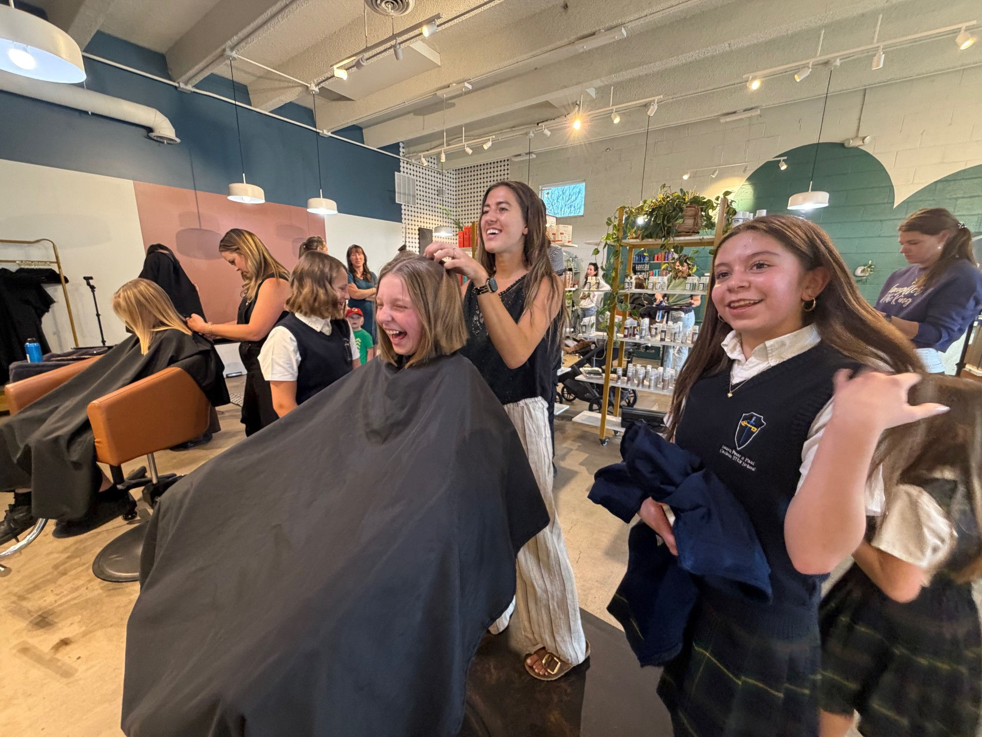 Group of girls and stylists gather in a salon, smiling and watching as one girl gets her hair cut.