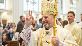 Bishop in ornate robes and mitre holds crosier, making peace sign. Background: church interior with stained glass, congregation smiling.