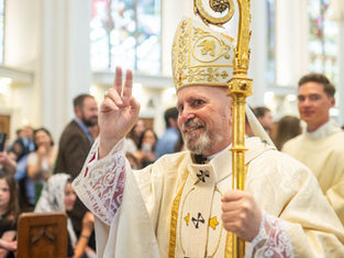 Bishop in ornate robes and mitre holds crosier, making peace sign. Background: church interior with stained glass, congregation smiling.
