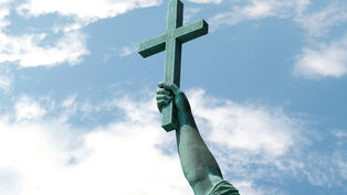 Bronze statue arm holding a cross aloft against a blue sky with scattered clouds