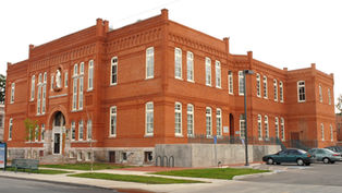 Red brick building with white-trimmed windows, featuring a statue above the entrance. Cars parked in front. Cloudy sky. Calm setting.