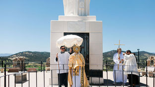 Priests conduct an outdoor Catholic ceremony at a white monument, holding a monstrance and canopy with mountains behind.