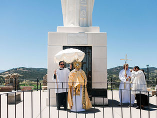 Priests conduct an outdoor Catholic ceremony at a white monument, holding a monstrance and canopy with mountains behind.