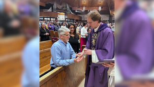Priest in purple robe blesses the hands of a man in blue at a church. Congregation stands in pews. Wooden ceiling and organ pipes visible.