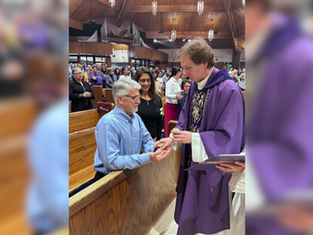 Priest in purple robe blesses the hands of a man in blue at a church. Congregation stands in pews. Wooden ceiling and organ pipes visible.