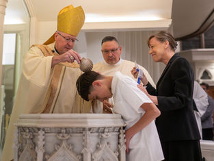Bishop pours water over a bowed boy during a baptism, while a smiling woman supports him in a church.