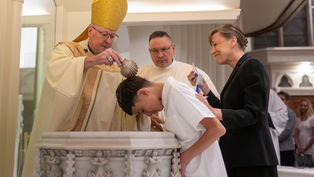 Bishop pours water over a bowed boy during a baptism, while a smiling woman supports him in a church.