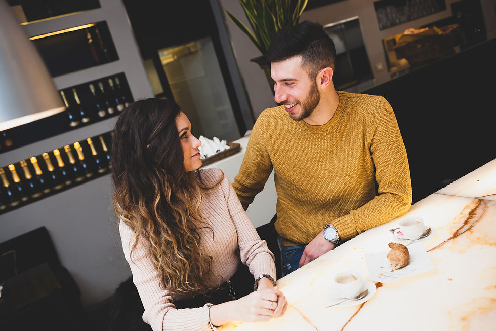 A couple sits at a cafe table, smiling at each other. He wears a mustard sweater, she a pink one. Cups of coffee and a pastry are on the table.