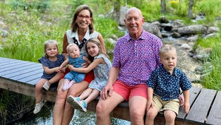 Smiling adults and children sit on a wooden bridge over a stream in a lush, green park. Bright and cheerful atmosphere.