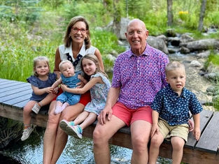 Smiling adults and children sit on a wooden bridge over a stream in a lush, green park. Bright and cheerful atmosphere.